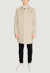 Trench coat impermeabile beige con colletto abbottonato, tasche frontali e polsini arrotolati, indossato sopra pantaloni neri slim-fit e sneakers bianche.