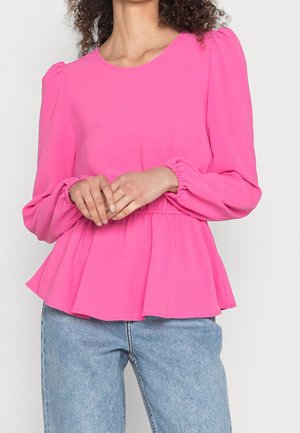 Femme portant un chemisier rose vif à manches longues avec une taille froncée et des manches bouffantes, associé à un jean bleu clair, mains jointes devant.