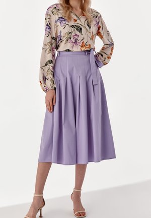 Femme portant un chemisier à manches longues à fleurs, rentré dans une jupe midi lavande taille haute avec des plis et une ceinture nouée sur le côté.