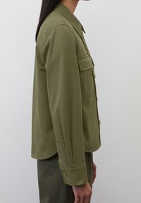 Camisa chaqueta de manga larga en verde oliva, con cuello puntiagudo, bolsillo en el pecho delantero y cierre de botones. Tejido suave, ajuste ligeramente entallado.