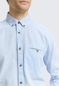 Lichtblauw button-up overhemd met een spread collar, bronzen knopen en een borstzak met een klep. De stof heeft een subtiele textuur.