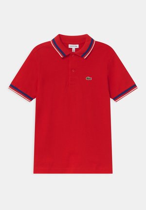 Červené polo tričko s krátkým rukávem, se modrým, bílým a červeným lemem na límci a rukávech, na hrudi má malé zelené logo krokodýla.