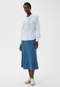 Donna in piedi con una blusa leggera blu a volant, una gonna midi in denim blu e scarpe da ginnastica bianche con strisce nere su uno sfondo neutro.