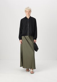 Svart zip-up bomberjakke med rund krage, kombinert med et langt olivengrønt satengskjørt og beige sandaler. Holder en svart håndveske.