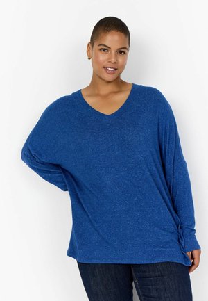 Plavi V-izrez oversized top napravljen od mekanog materijala s teksturiranim izgledom. Ima rolane rukave i čvor na rubu.