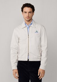 Hombre vistiendo una chaqueta beige con cremallera sobre una camisa azul clara, de pie contra un fondo gris liso, mirando hacia adelante.