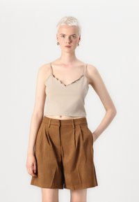 Crop top beige à bords festonnés avec des fines bretelles, associé à un short plissé marron taille haute. Textures de tissu lisses avec une coupe décontractée.