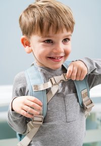Mochila azul claro con correas beige, que presenta una hebilla de plástico y tela texturizada. Niño ajustando la hebilla, vistiendo una camiseta gris de canalé.