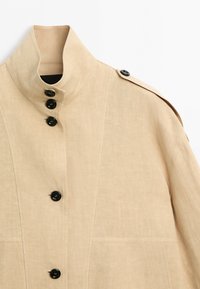 Beige linen jacket with stand-up collar, black buttons down front and on shoulder epaulet.