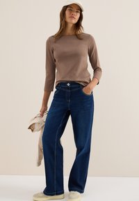 Top marrón de manga larga, jeans oscuros de cintura alta con bolsillos delanteros, zapatillas de color claro, sosteniendo una chaqueta beige. Fondo neutro.