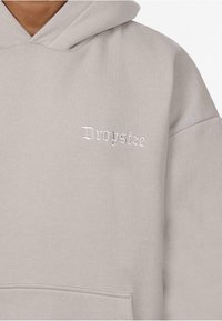 Helle graue Kapuzenjacke aus einem Stoff mit weicher Textur. Verfügt über eine Vordertasche und ein aufgesticktes "Dropsize"-Logo in Weiß. Verstellbare Kapuze.