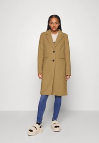 Płaszcz z mieszanki wełny w oliwkowej zieleni z wyciętym kołnierzem, dwoma guzikami i bocznymi kieszeniami, zestawiony z niebieskimi dżinsami i beżowymi pluszowymi sandałami.