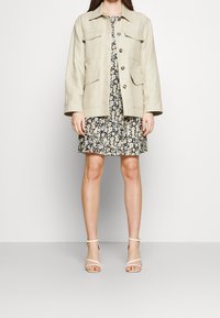 Veste beige à col avec cinq poches, boutonnée, portée par-dessus une robe noire à motifs floraux avec une jupe plissée. Sandales blanches à brides.