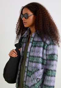 Purple and green plaid jacket with fuzzy texture, worn over an olive green outfit, holding a black quilted shoulder bag, accessorized with round black sunglasses.