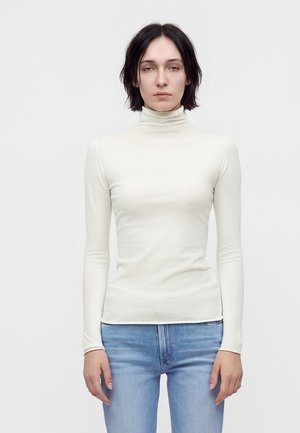 Mujer con el cabello corto y oscuro, vistiendo un jersey de cuello alto blanco y vaqueros azul claro, de pie contra un fondo claro y liso.
