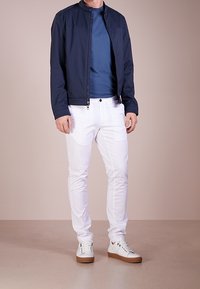Marinejakke med høj krave, blå skjorte, hvide bukser og hvide sneakers med brune såler. Casual outfit vist på en neutral baggrund.