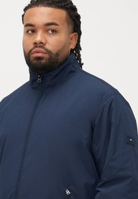 Geacă bleumarin cu fermoar și guler înalt, dintr-un material neted. Dispune de o etichetă cu logo pe mâneca stângă și buzunar lateral cu fermoar.