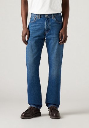 Man draagt blauwe denim jeans met rechte pijpen, gecombineerd met donkerbruine veterschoenen en een wit overhemd.