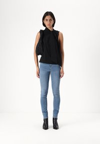 Top preto sem mangas com detalhe de nó na frente, combinado com jeans justos azuis e botas pretas de cano curto, segurando uma bolsa texturizada preta.
