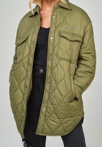 Chaqueta acolchada de color verde oliva con botones a presión, dos bolsillos delanteros y un acabado suave al tacto, llevada sobre una camiseta negra y jeans oscuros.