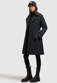 Trench coat impermeabile nero con bottoni doppi, tasche laterali e polsini regolabili, abbinato a leggings neri e stivali neri robusti.