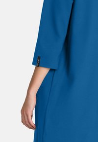 Tunic albastră cu mâneci trei sferturi, având o textură netedă și un etichetă mică neagră cu text alb aproape de mansetă.