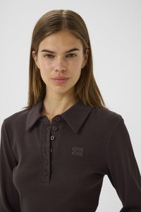 Young woman with straight brown hair wearing a dark brown ribbed long-sleeve polo shirt with buttons and a small embroidered logo on chest.