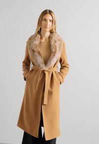 Palton din camel cu guler din blană sintetică, talie accentuată cu cordon și mâneci lungi. Textură netedă, design până la genunchi, și fante laterale pentru o mișcare ușoară.
