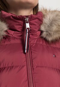 Bordowa puchowa kurtka z kapturem wyściełanym futrem, wyposażona w pełny zamek błyskawiczny oraz ozdobną tasiemkę w paski. Uwaga: logo widoczne na lewej piersi.