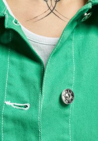 Primer plano de una chaqueta verde con un botón de metal etiquetado "ONLY", llevada sobre una camiseta blanca de canalé, mostrando una zona de cuello tatuada arriba.