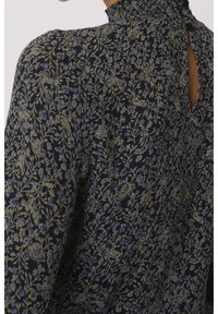 Blouse foncée avec motif floral et oiseau beige et bleu, col montant froncé et petite ouverture en forme de trou de serrure au dos avec fermeture bouton.