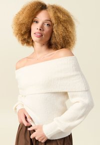 Sweter z odkrytymi ramionami z prążkowanej dzianiny w kolorze kremowym, z długimi rękawami i przyjemną fakturą. Modelka ma na sobie zwiewną brązową spódnicę. Neutralne tło.