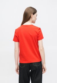 Κόκκινο βαμβακερό κοντομάνικο μπλουζάκι με στρογγυλή λαιμόκοψη και άνετη εφαρμογή. Η πίσω όψη δείχνει ομοιόμορφη υφή και απλό σχέδιο. Φορεμένο με σκούρα τζιν.