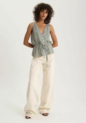 Femme aux cheveux bouclés porte un top sans manches à motifs avec boutons et ceinture à nouer à la taille, associé à un pantalon large crème et des sandales noires.