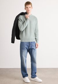 Hellgrüner, gerippter Pullover, schwarze Jacke über der Schulter drapiert, blaue Jeans, weiße Sneaker, minimalistischer Stil, glatte Textur, lässige Passform.