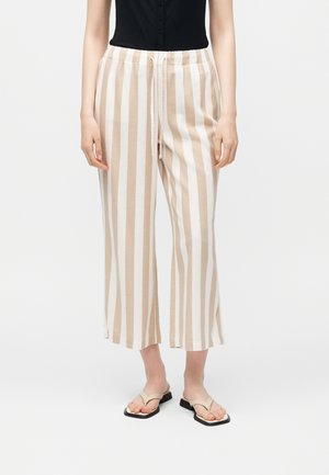 Vrouw die beige en witte verticale gestreepte wijde cropped broek met trekkoord in de taille en beige teenslippers draagt, staand tegen een witte achtergrond.