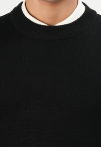 Close-up van een persoon die een zwart gebreid sweater met ronde hals draagt over een witte shirtkraag die aan de nek zichtbaar is.