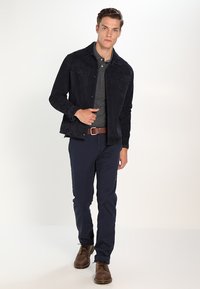 Veste en daim marine sur un polo gris, assortie à un pantalon marine et des bottes marron. Comprend des poches poitrine et une fermeture à boutons.