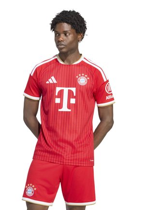 Mand stående iført rød FC Bayern München fodboldtrøje og shorts, kiggende til siden med hænderne bag ryggen.