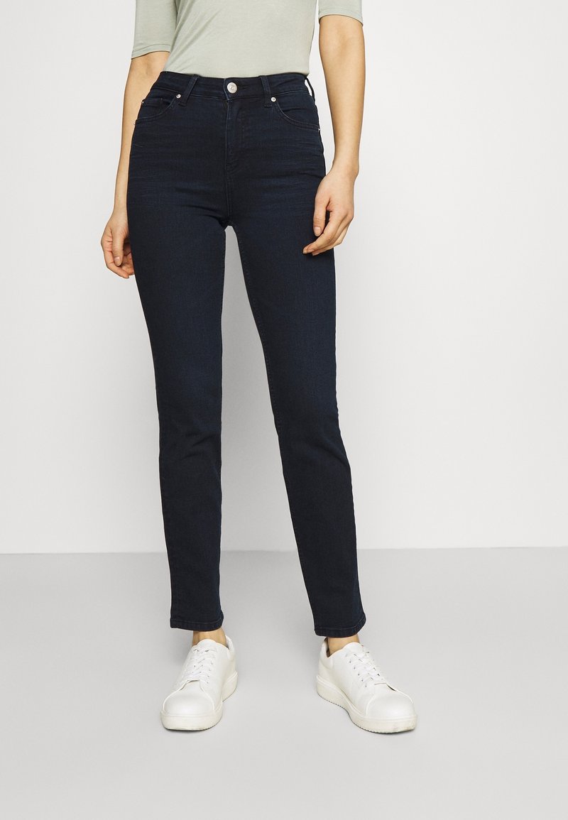 Jeans de mezclilla oscuro con un ajuste slim, cintura alta, diseño de cinco bolsillos y cierre frontal con un solo botón metálico, combinados con zapatillas blancas.