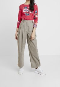 Rotes Blütenmuster Langarmoberteil, kombiniert mit hellbeigen hochtaillierten weiten Hosen. Modell trägt weiße Sneaker. Einheitlicher Hintergrund.