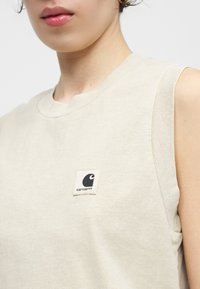 Κοντινό πλάνο ενός ατόμου που φοράει μπεζ αμάνικη μπλούζα με μικρό λογότυπο Carhartt στο στήθος.