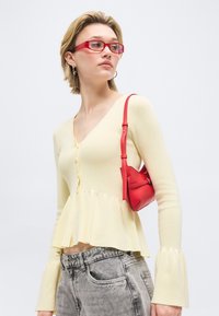 Νεαρή γυναίκα που φορά κόκκινα γυαλιά και ένα ανοιχτό κίτρινο πουκάμισο με κουμπιά και μανίκια που φουσκώνουν, κρατώντας μια μικρή κόκκινη τσάντα ώμου.