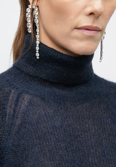 Marineblauer Strickrollpullover mit transparenter Textur, kombiniert mit langen, hängenden Kristallohrhängern mit mehreren rechteckigen Facetten.