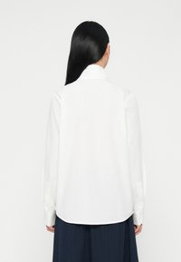 Camicia bianca a maniche lunghe realizzata in tessuto morbido, con colletto classico, polsini abbottonati e orlo curvo. Nessun motivo o accento.
