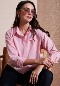 Striped pink and white button-up shirt with long sleeves and a chest pocket, worn with dark jeans. Accessories include oversized sunglasses.