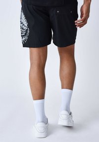 Bañador negro con estampado gráfico blanco en la pierna izquierda, que presenta dos bolsillos traseros con detalles de ojales, combinado con calcetines blancos y zapatillas.
