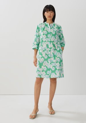 Floralkleid aus grünem Stoff mit weißen und hellblauen Blumen. Hat eine Knopfleiste, kurze Ärmel und Seitentaschen.