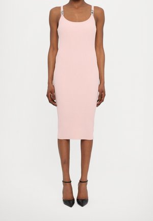 Kobieta ubrana w różową prążkowaną sukienkę midi na cienkich ramiączkach oraz czarne szpilki z zaostrzonym noskiem, stojąca na tle jednolitej białej ściany.