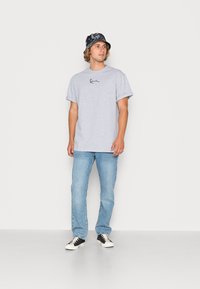 Grå bomullströja med korta ärmar och en svart logotyp, tillsammans med ljusblå jeans och en svart och grå mönstrad bucket-hatt.